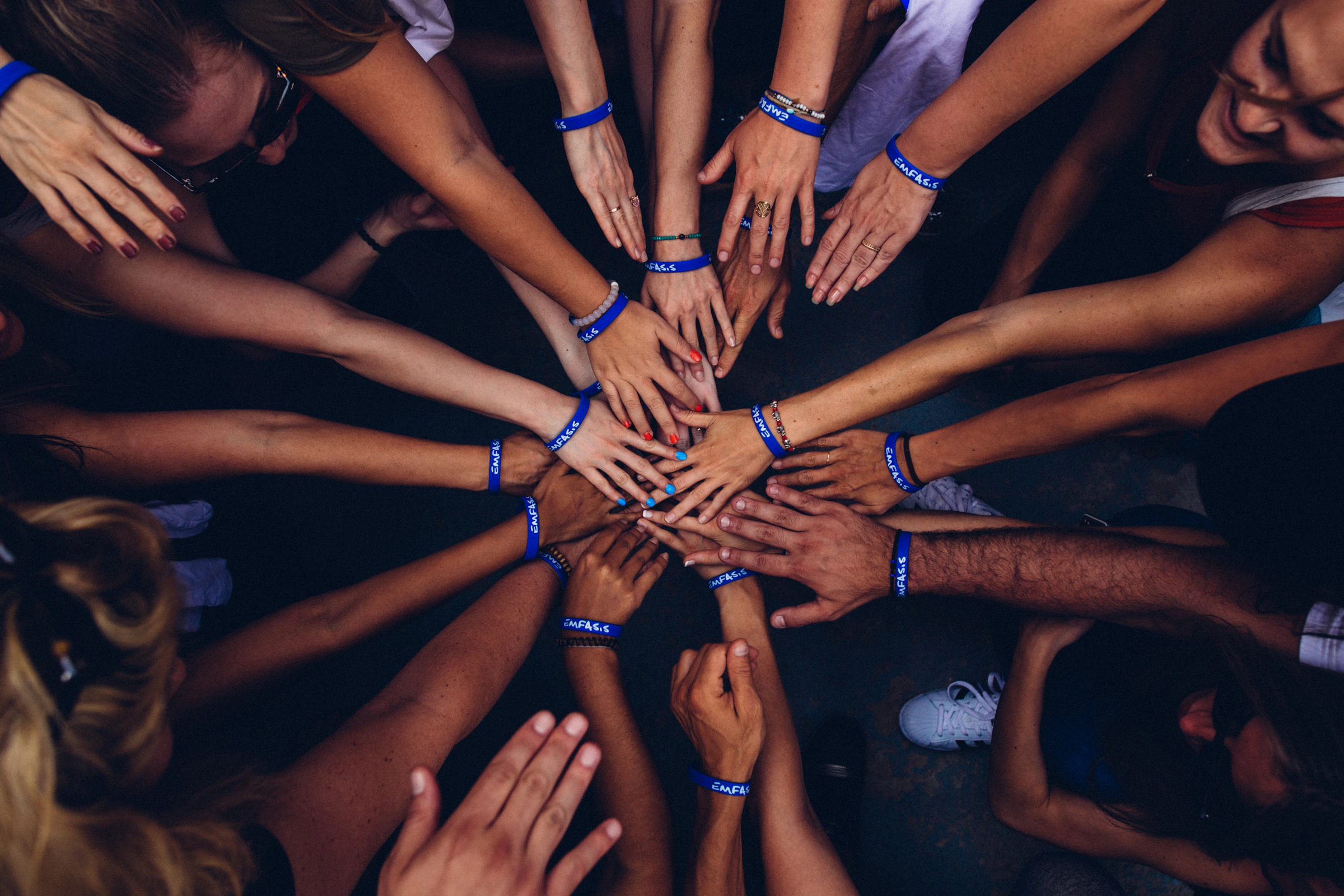An overhead view of a dozen people's hands and forearms reaching in from every direction, wrists banded, fingertips meeting at the center of a huddle.
